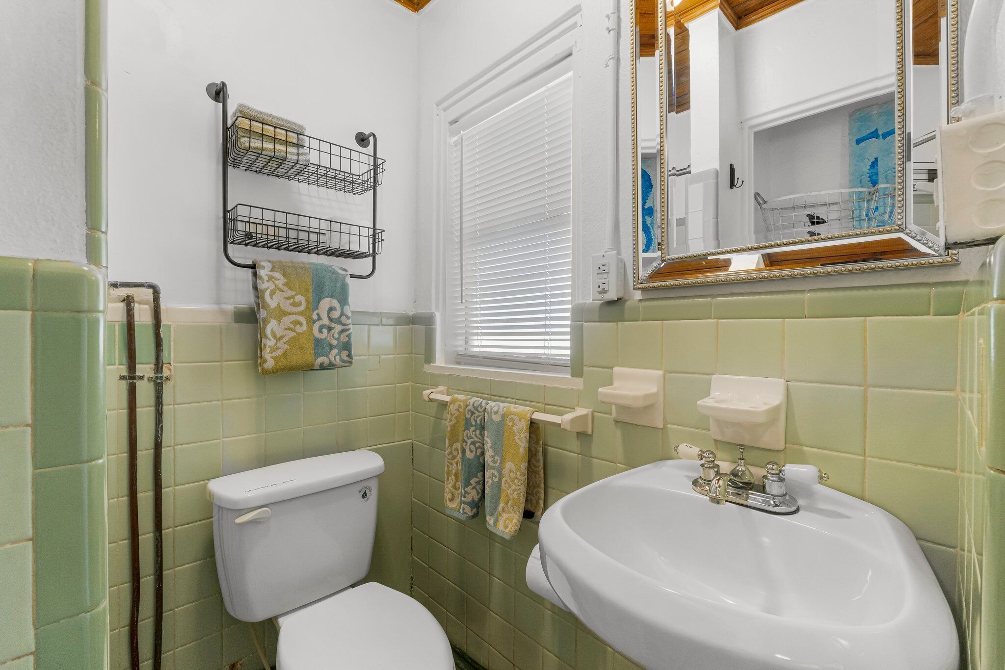 Une salle de bain équipée de toilettes, d’un lavabo et d’une fenêtre avec des stores. Des serviettes sont suspendues sur un porte-serviettes et un miroir est placé au-dessus du lavabo.
