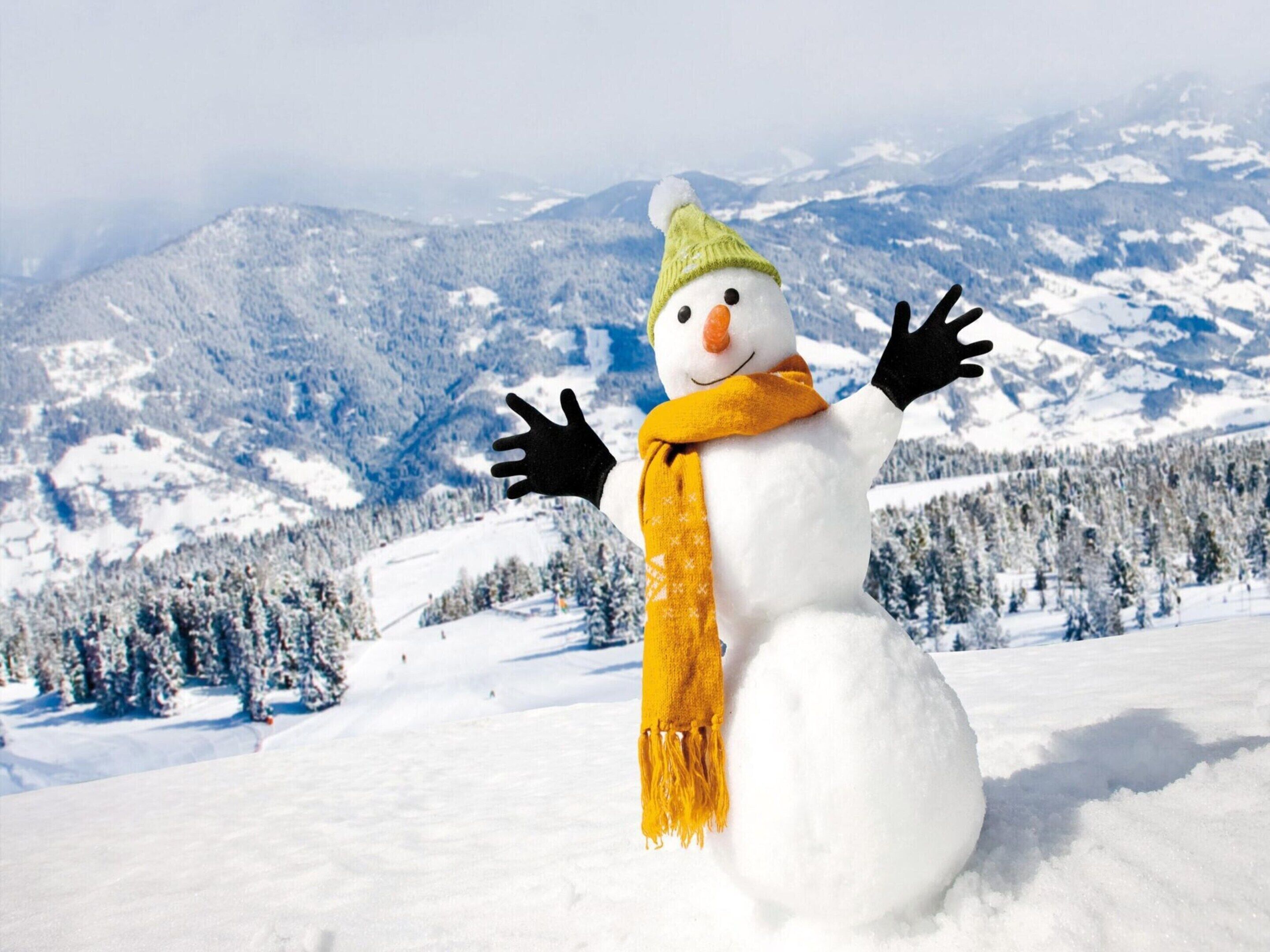 A snowman with a green hat and yellow scarf in a snowy landscape with mountains in the background.