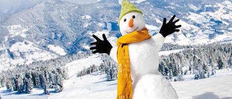 A snowman with a green hat and yellow scarf in a snowy landscape with mountains in the background.