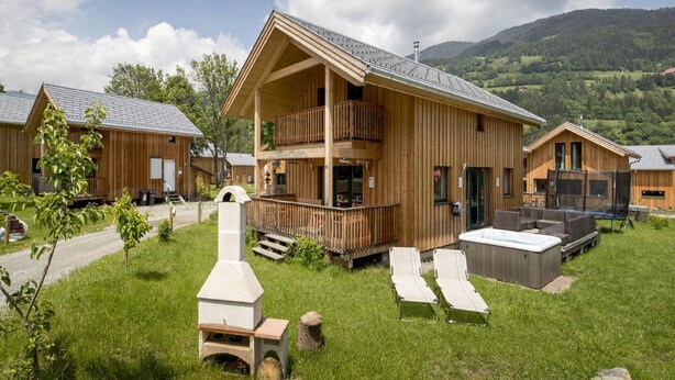 A wooden chalet with a balcony, a hot tub, and a stone oven in a grassy area with mountains in the background. - Pleasant Chalet With Indoor hot tub (Sankt Georgen am Kreischberg)