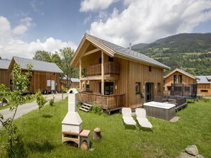 A wooden chalet with a balcony, a hot tub, and a stone oven in a grassy area with mountains in the background. - Pleasant Chalet With Indoor hot tub (Sankt Georgen am Kreischberg)