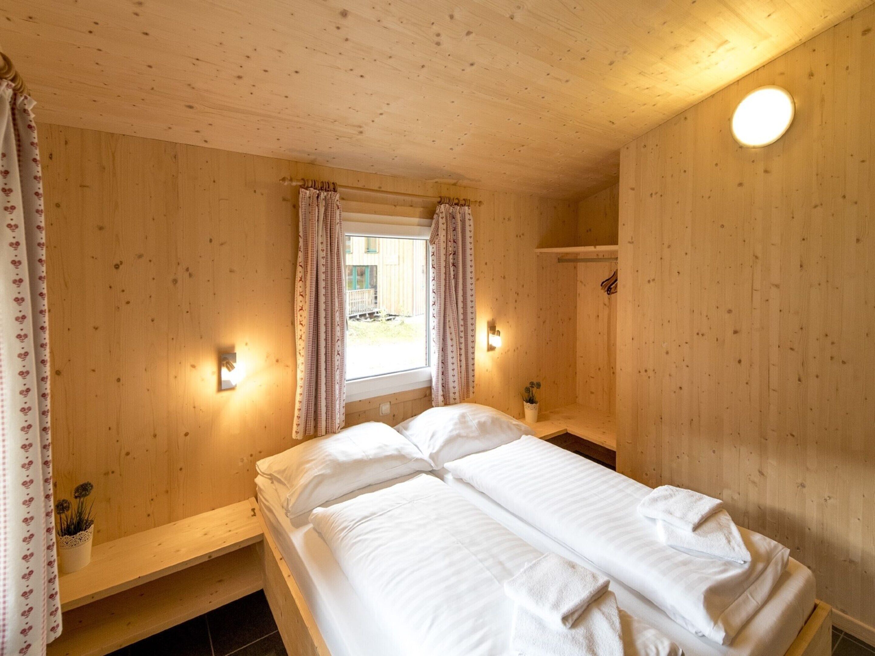 Una habitación de madera con cama, una ventana con cortinas, un estante pequeño y una lámpara fijada a la pared.
