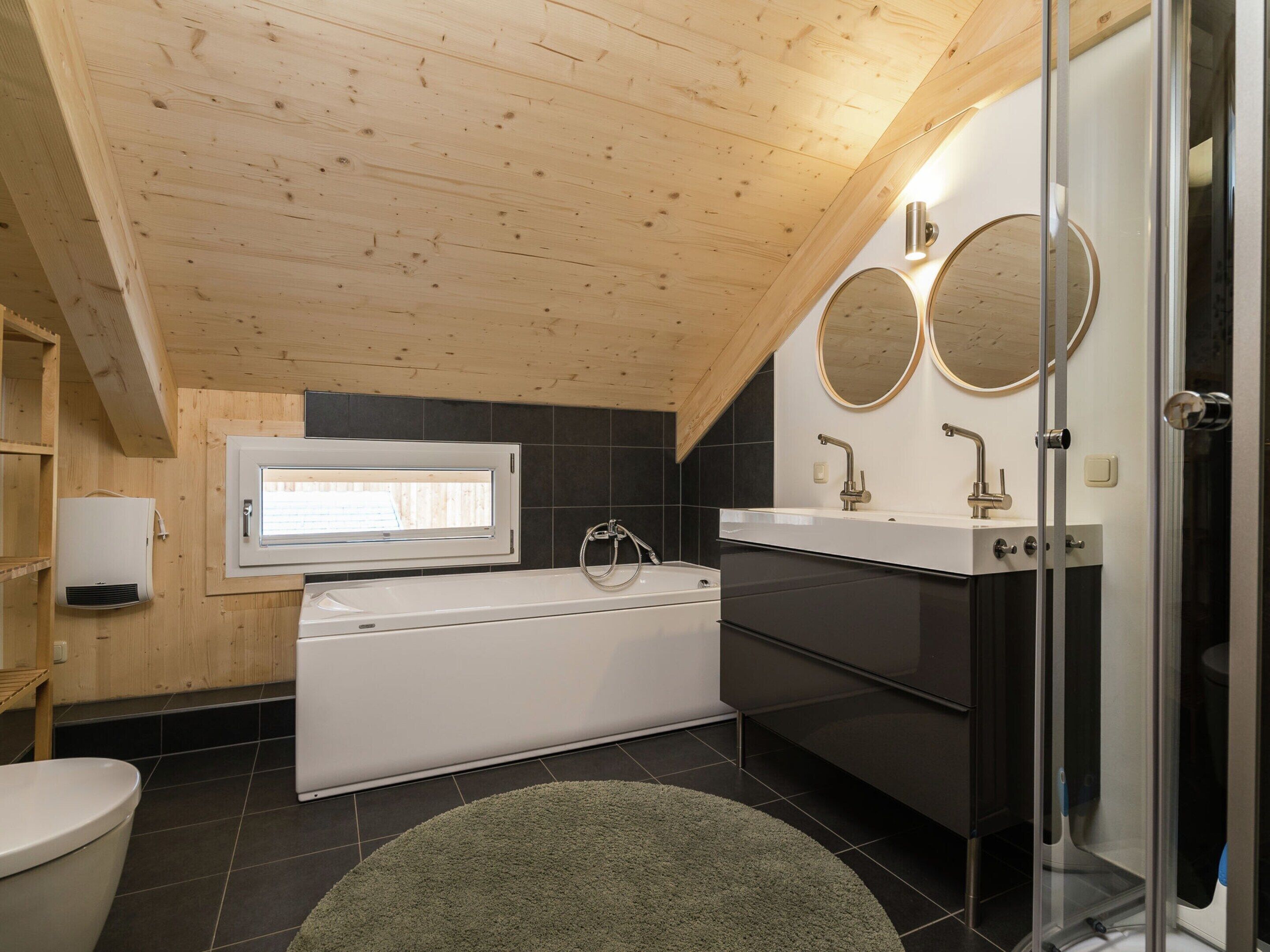 A modern bathroom with a bathtub, a shower, and a toilet. There is a window above the bathtub and two round mirrors above the sink.