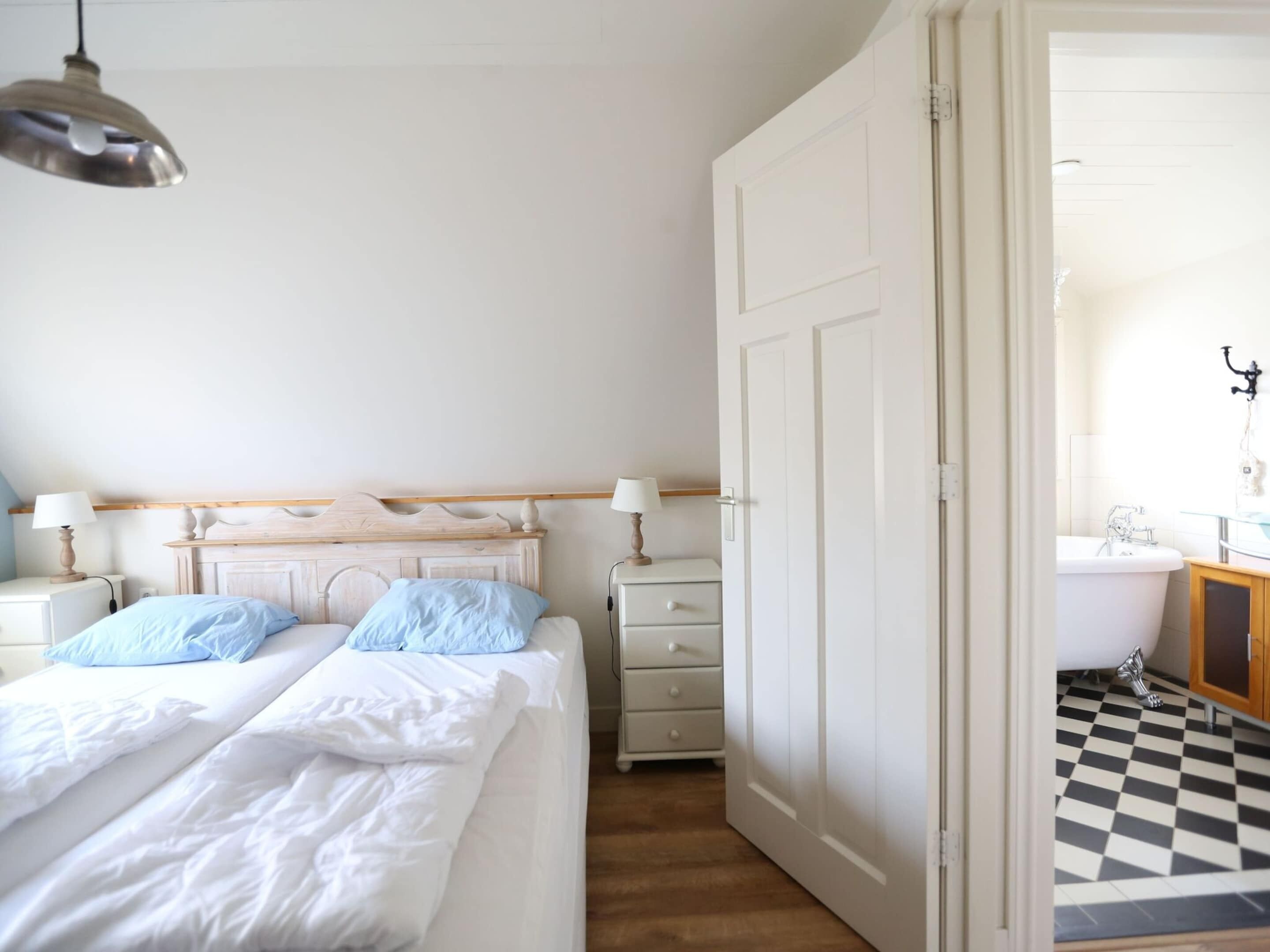 A bedroom with a bed, bedside tables, a nightstand, a lamp, and a door leading to a bathroom with a checkered floor.