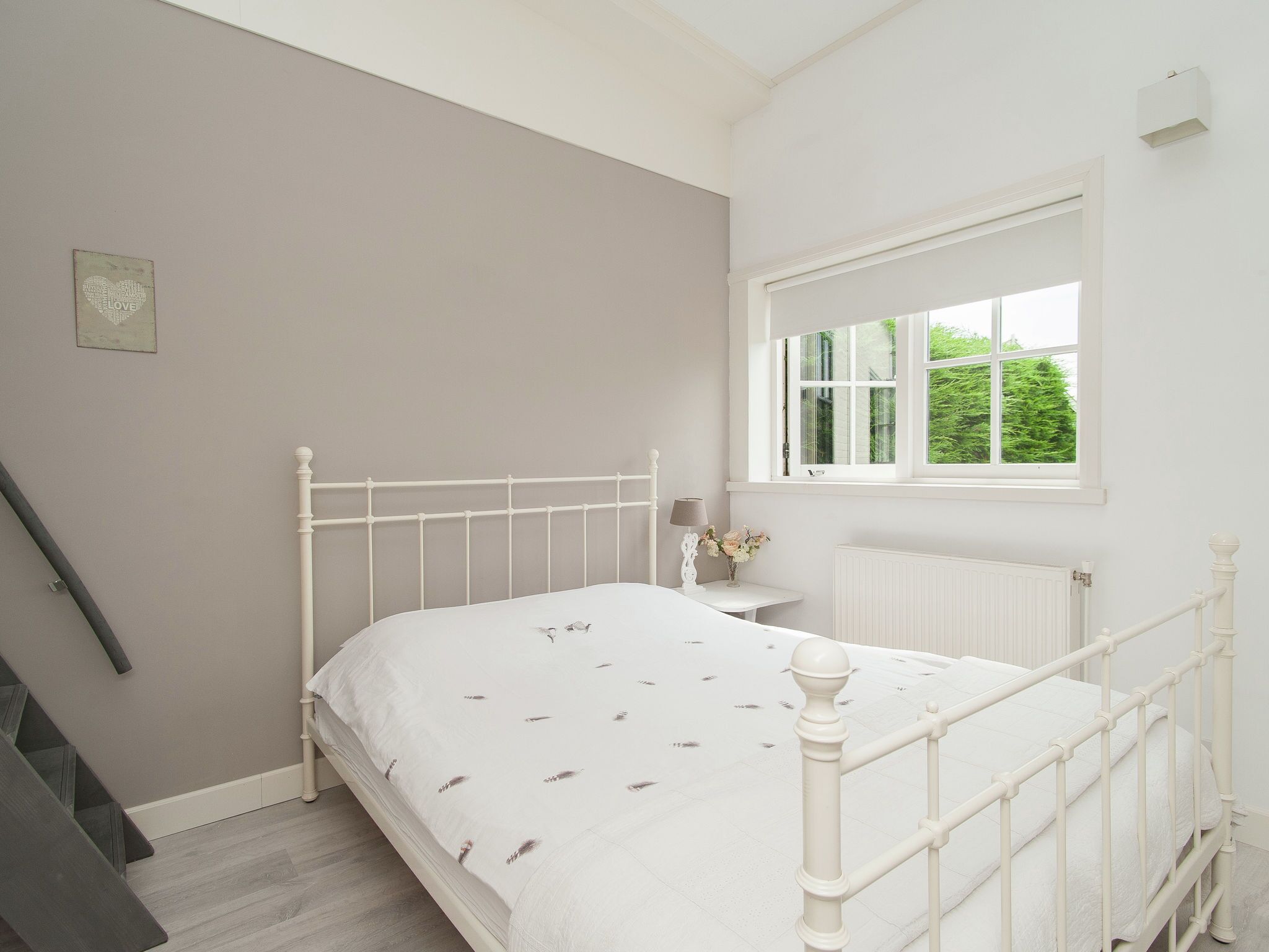 Une chambre avec un lit blanc, une petite table de chevet avec une lampe, une fenêtre donnant sur un paysage verdoyant et un radiateur.