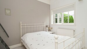 A bedroom with a white bed, a small bedside table with a lamp, a window with a view of greenery, and a radiator. - Holiday Home Egmond Near Dune Beaches (Egmond-Binnen)