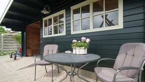 An outdoor patio with a round metal table and two cushioned chairs, a potted plant with flowers, and a wooden fence in the background. - Holiday Home Egmond Near Dune Beaches (Egmond-Binnen)