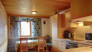 Eine kleine Küche mit Holzausstattung, einem Esstisch mit Stühlen, einem Fenster mit gemusterten Vorhängen und einem Fernseher.