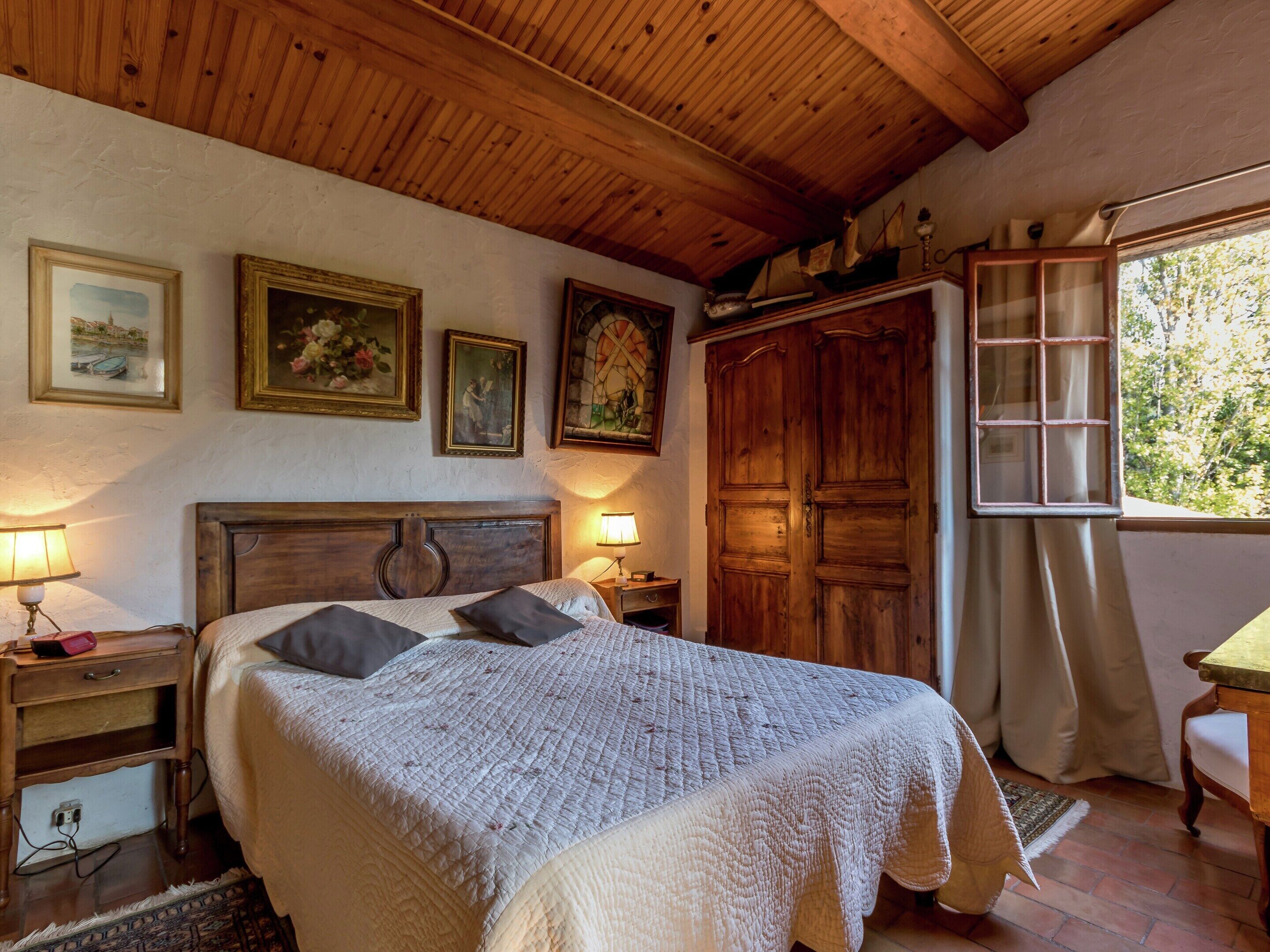 Un dormitorio con techo de madera, una cama con colcha blanca, una mesita de noche con lámpara, un armario y una ventana con vistas a árboles.