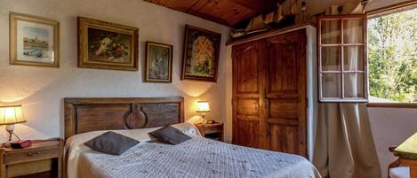 Un dormitorio con techo de madera, una cama con colcha blanca, una mesita de noche con lámpara, un armario y una ventana con vistas a árboles.