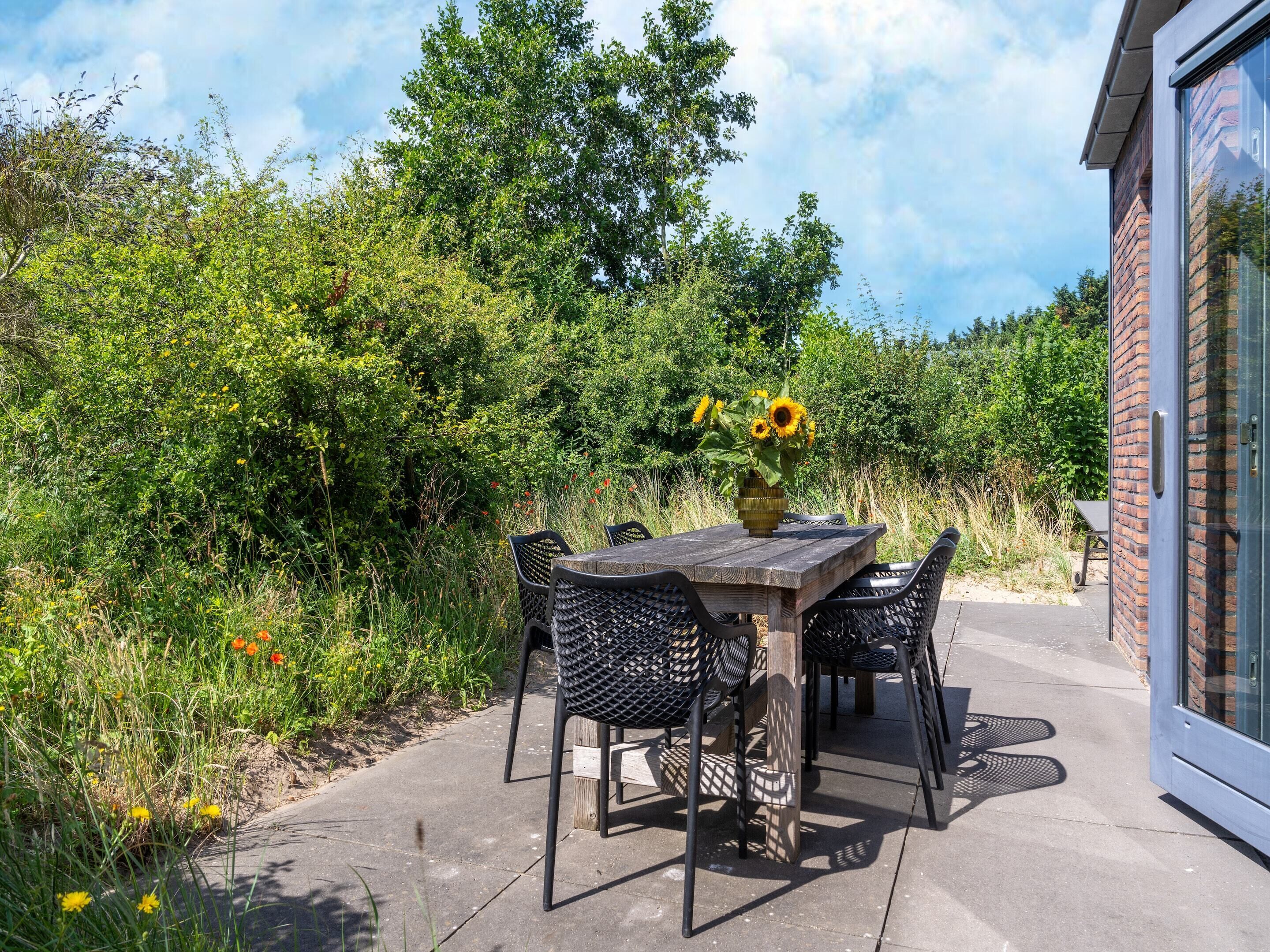 An outdoor patio with a wooden table and chairs, surrounded by lush greenery and a brick building.