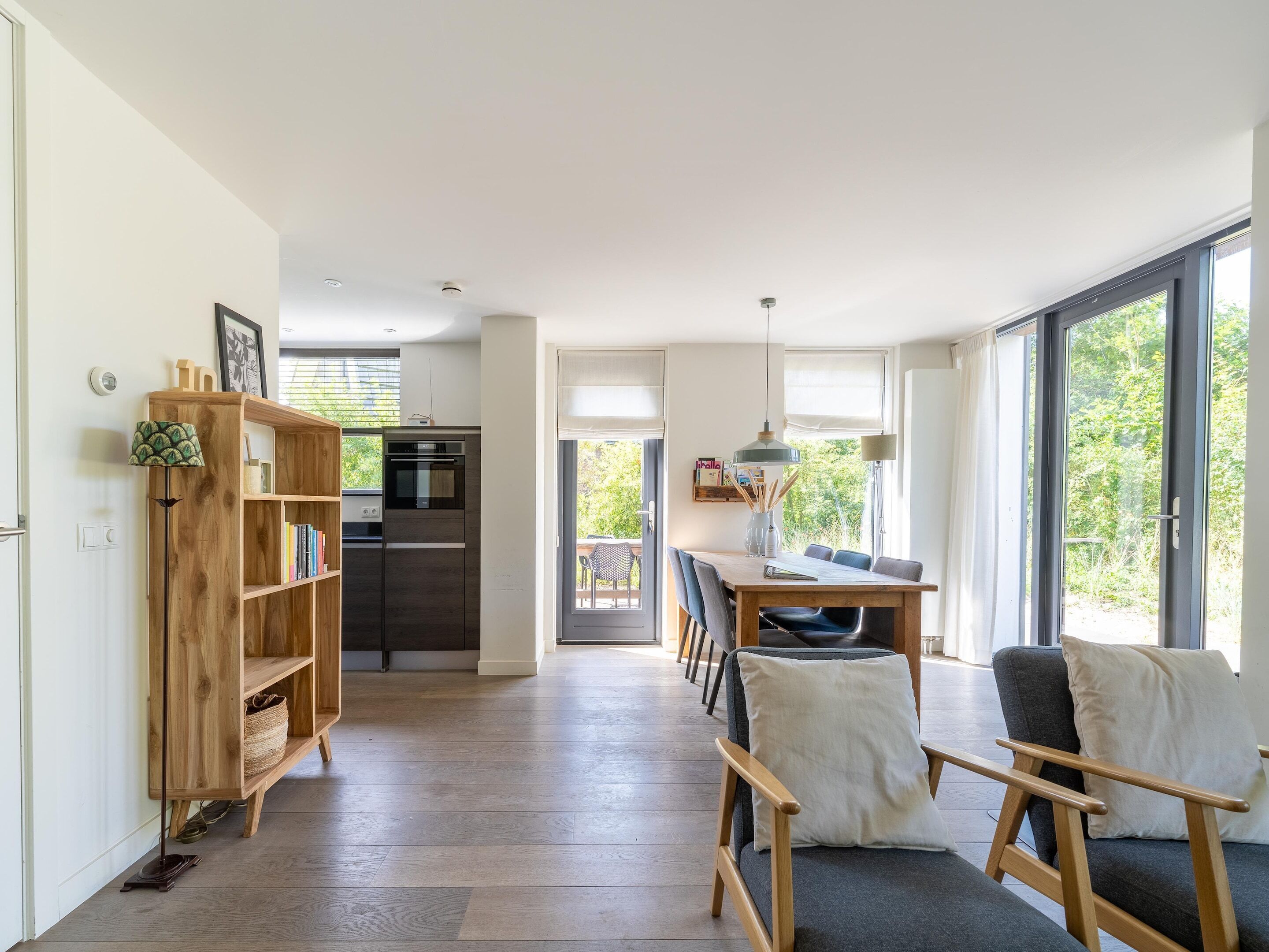 A spacious living area with a wooden bookshelf, a dining table with chairs, and a kitchen in the background.