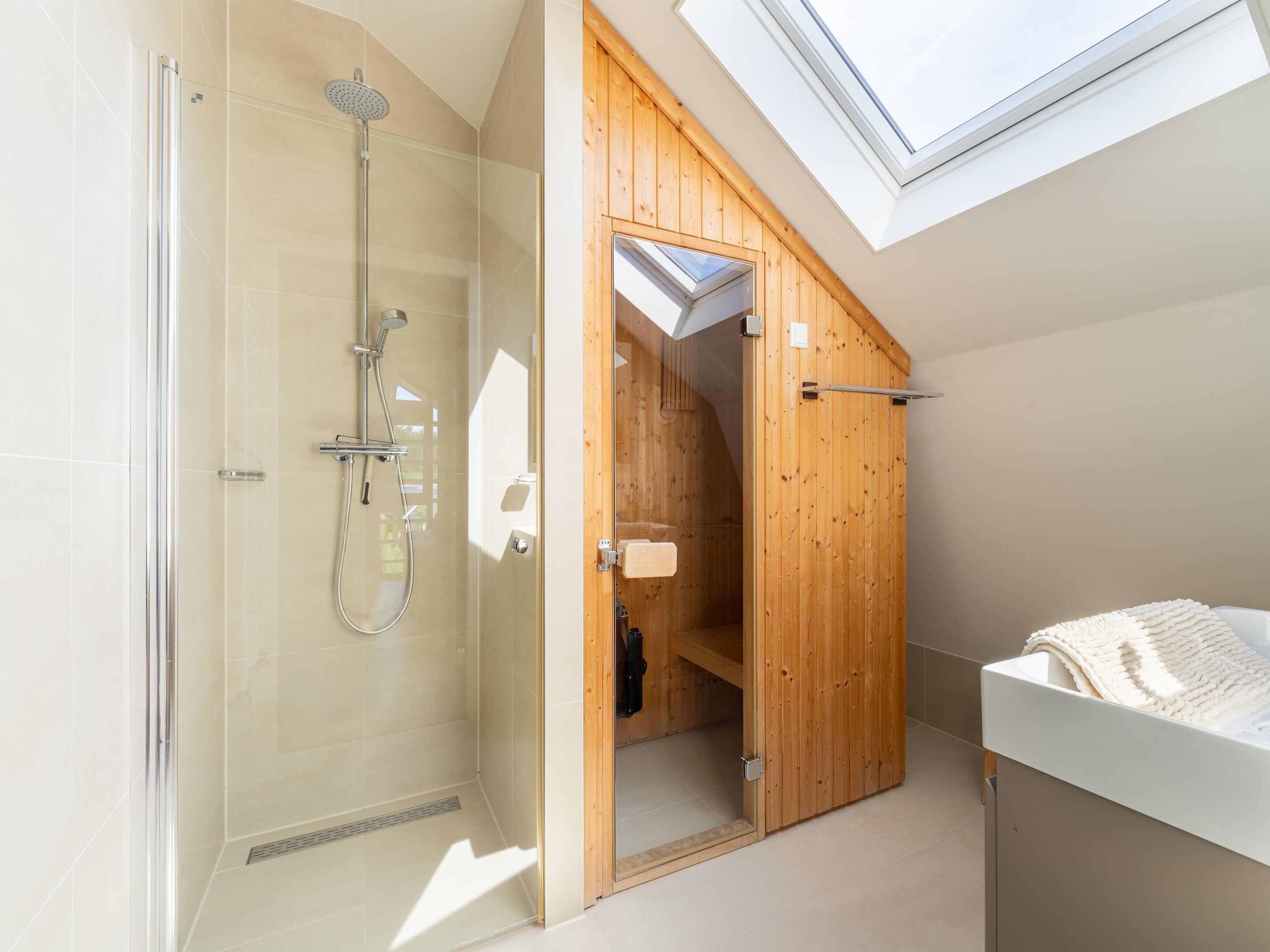 A modern bathroom with a glass-enclosed shower, wooden door, skylight, and a white sink.