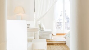 A bright, white room with a bed, a small table with a lamp, and a view of buildings through sheer curtains.