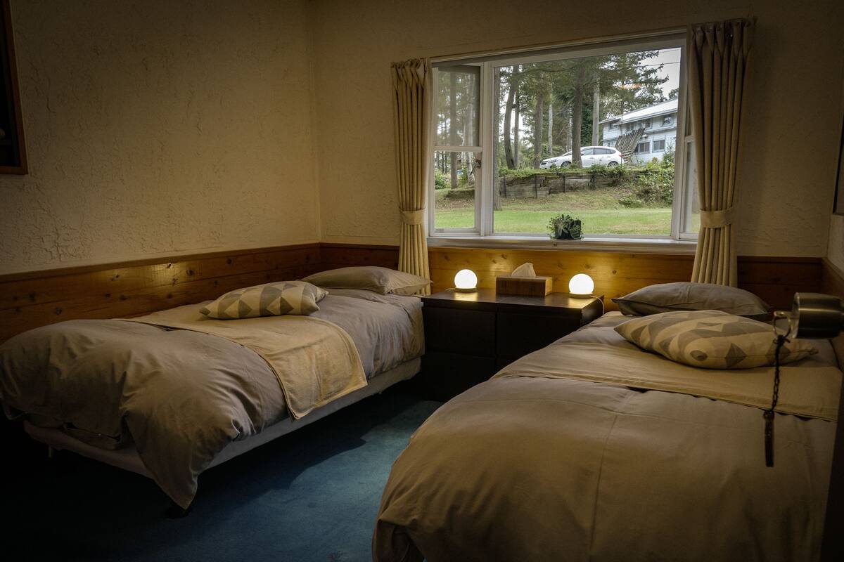 A room with two beds, a window with curtains, a bedside table with a lamp, and a view of a garden and parked car outside.