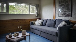 A room with a grey sofa, a small wooden table with a cushion, and a window with curtains. - Japan Snowsports (Myoko)
