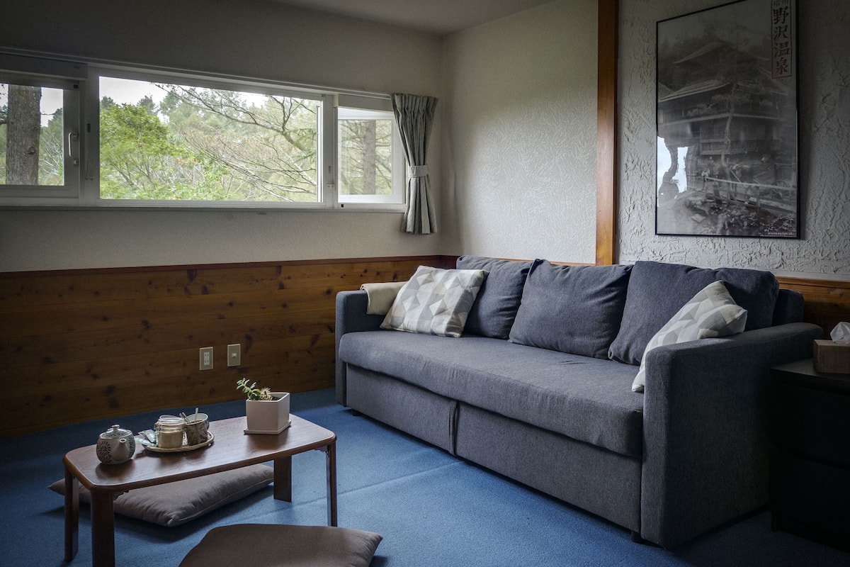 A room with a grey sofa, a small wooden table with a cushion, and a window with curtains.