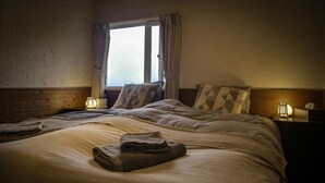 A neatly made bed with a blanket and a folded towel, a bedside lamp, and a window with curtains. - Japan Snowsports (Myoko)