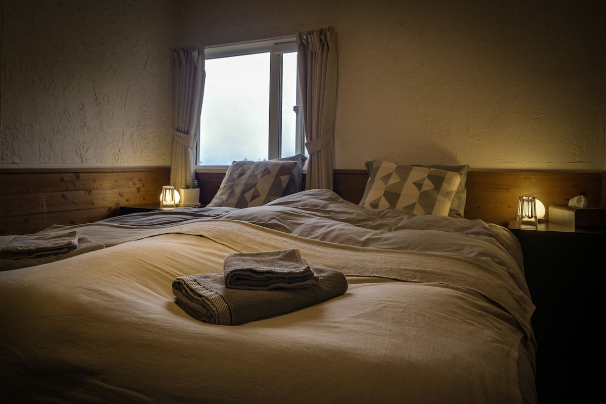 A neatly made bed with a blanket and a folded towel, a bedside lamp, and a window with curtains.