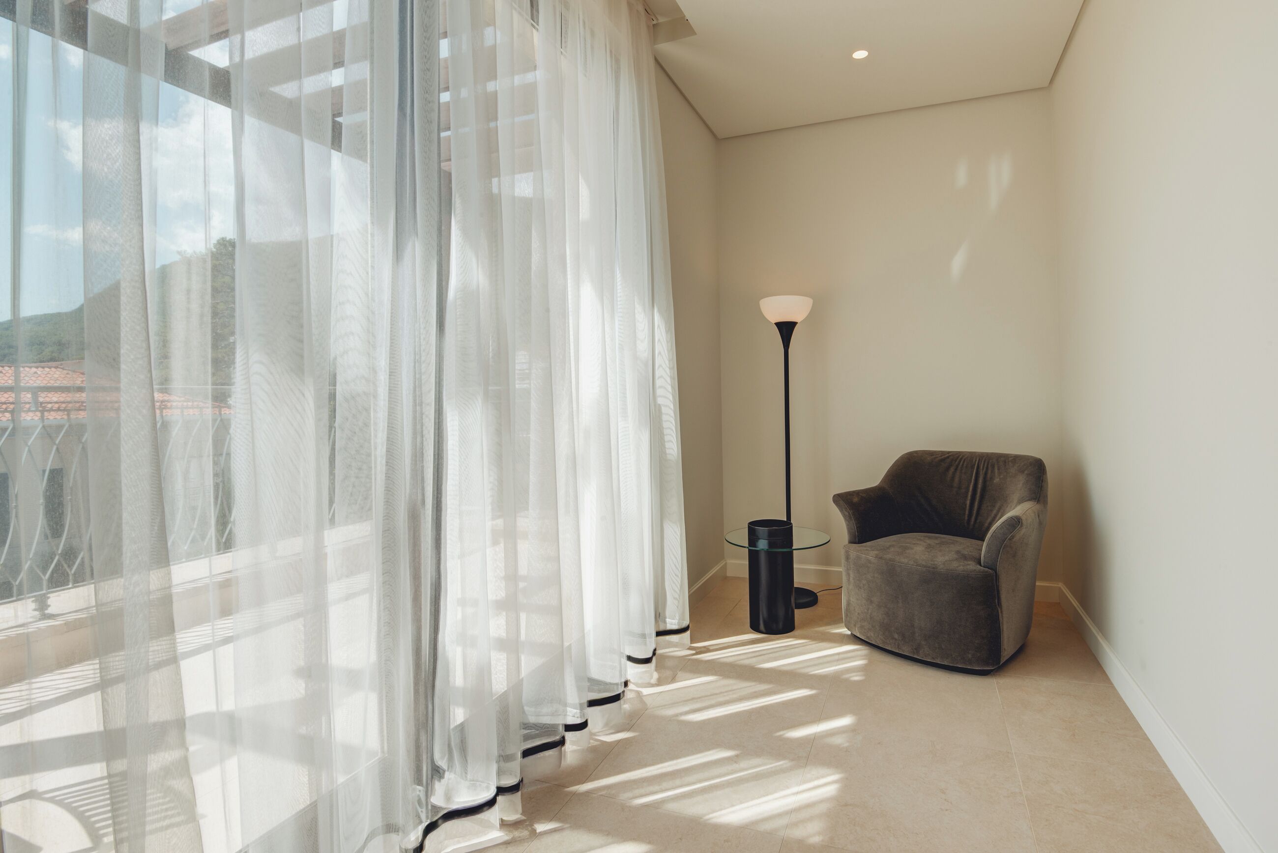 Habitación de esquina con cortinas traslúcidas, una lámpara de pie y un sillón gris.