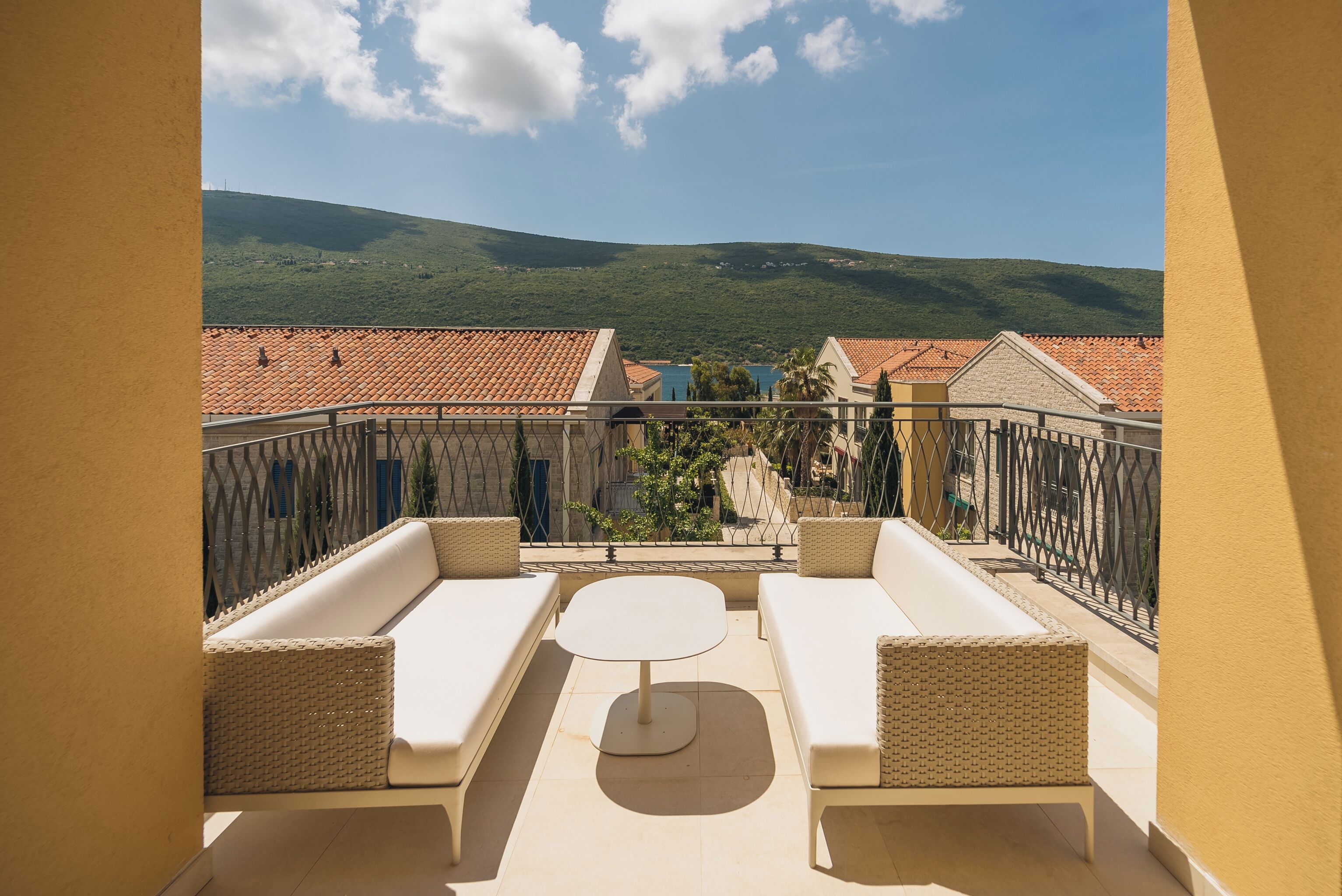 A balcony with two white sofas and a round table, overlooking a hillside with terracotta-roofed buildings.