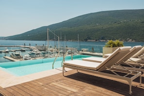 A poolside area with lounge chairs, a wooden deck, and a view of boats and a hill in the background.