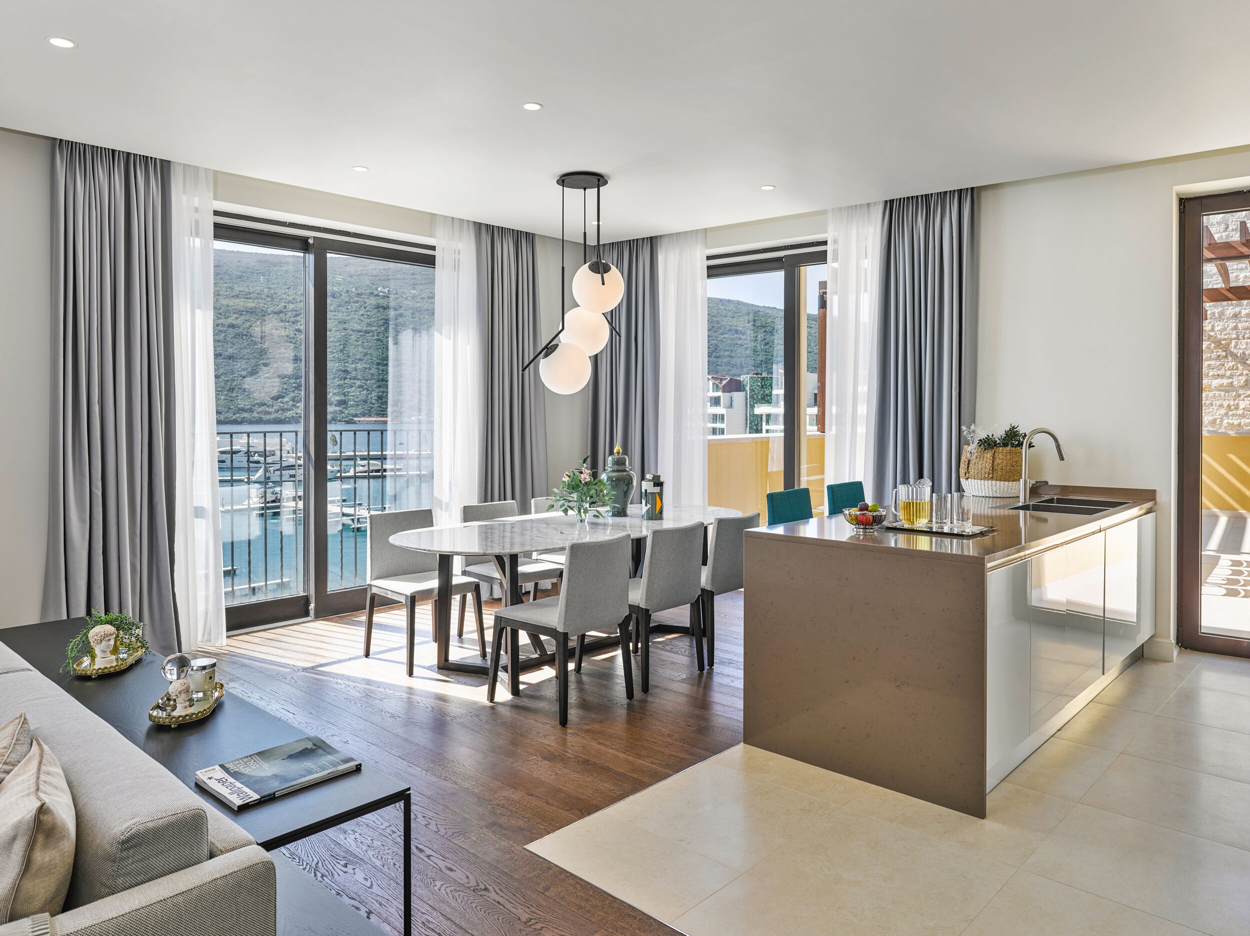 Un apartamento moderno con comedor, cocina y grandes ventanales que ofrecen vistas al mar y a las colinas.
