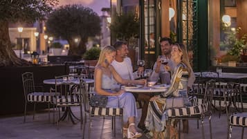Een terras met ronde tafels en stoelen, omgeven door groen en lantaarns.