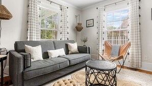 Ein Wohnzimmer mit grauem Sofa, einem runden Couchtisch und einem gewebten Hocker. Zwei Fenster mit Streifenmustervorhängen, eine Pendelleuchte und ein gerahmtes Bild an der Wand sind zu sehen.