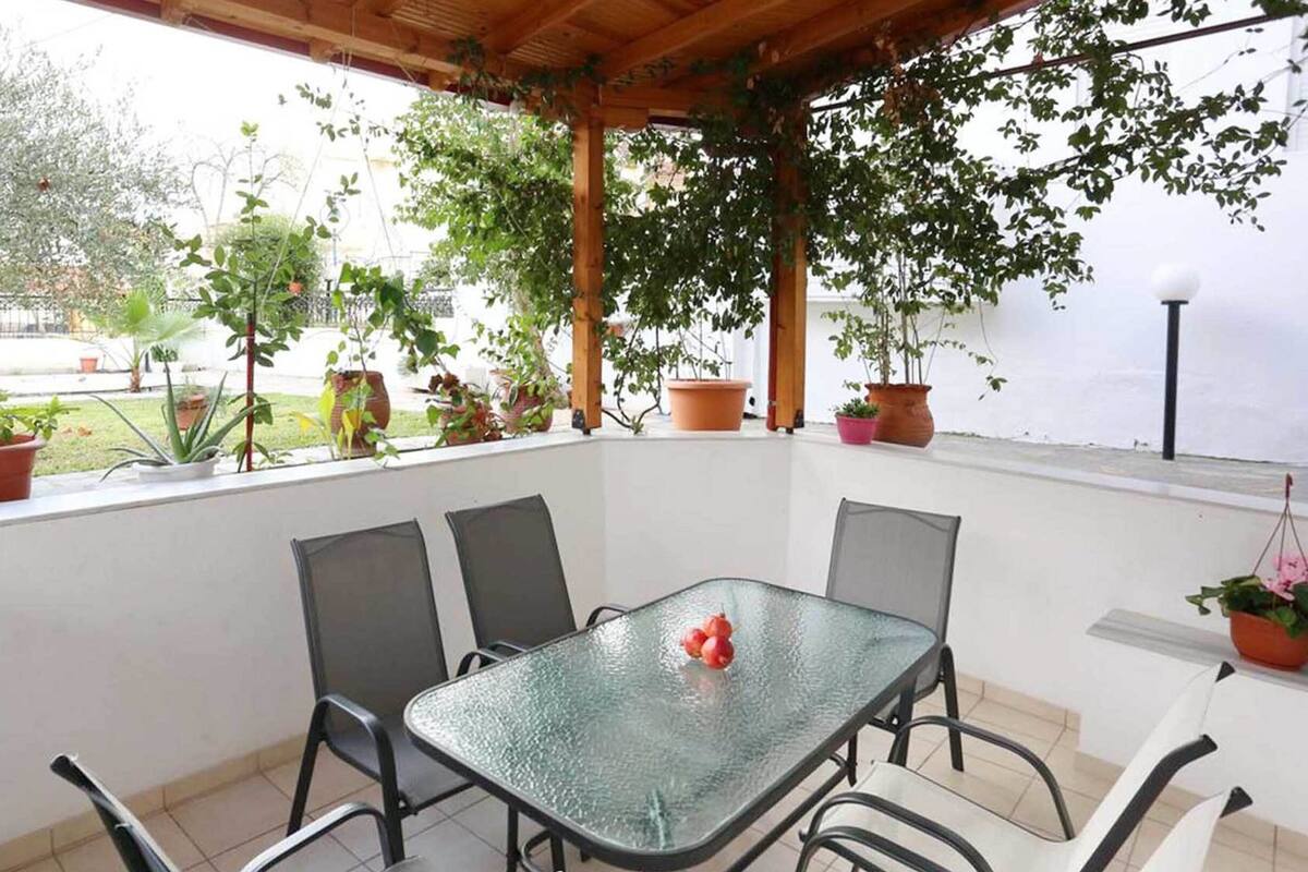 A covered patio with a glass table and chairs, surrounded by potted plants and a view of a garden.