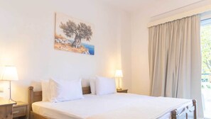 A hotel room with a bed, two bedside lamps, a painting of a tree on the wall, and a large window with curtains. - Anastasia Studios (Kassandra)