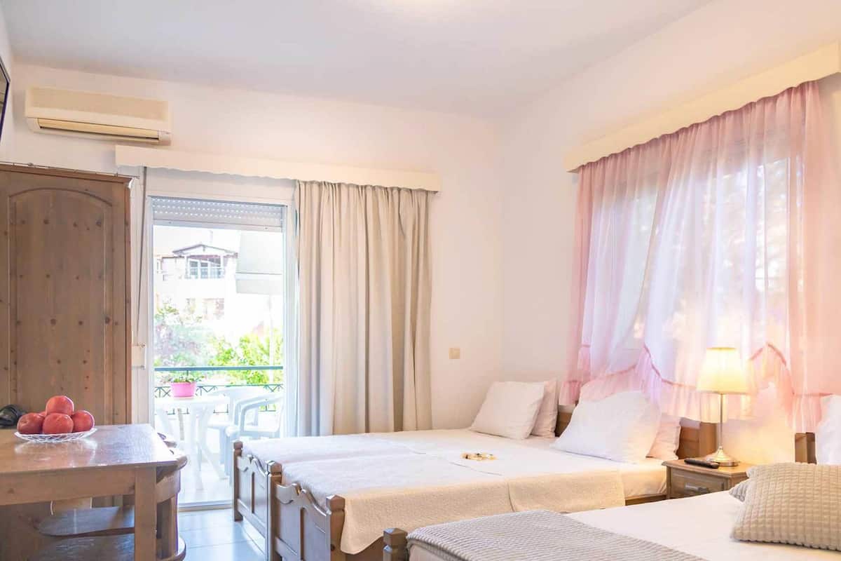 A hotel room with two beds, a desk with a basket of fruit, a table, and a balcony with a view of greenery and a building.