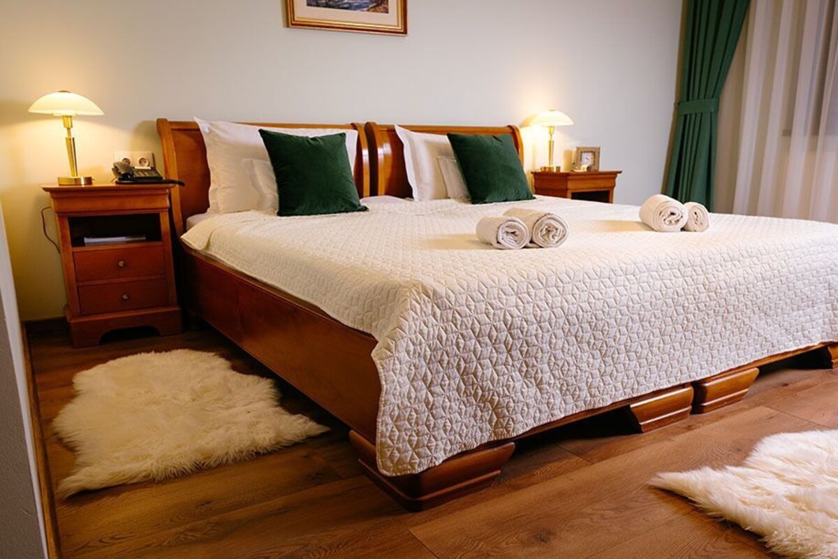 A neatly made bed with a quilted cover, two green pillows, and rolled towels on the bed. There are two bedside tables with lamps, a wooden headboard, and a fluffy rug on the floor.