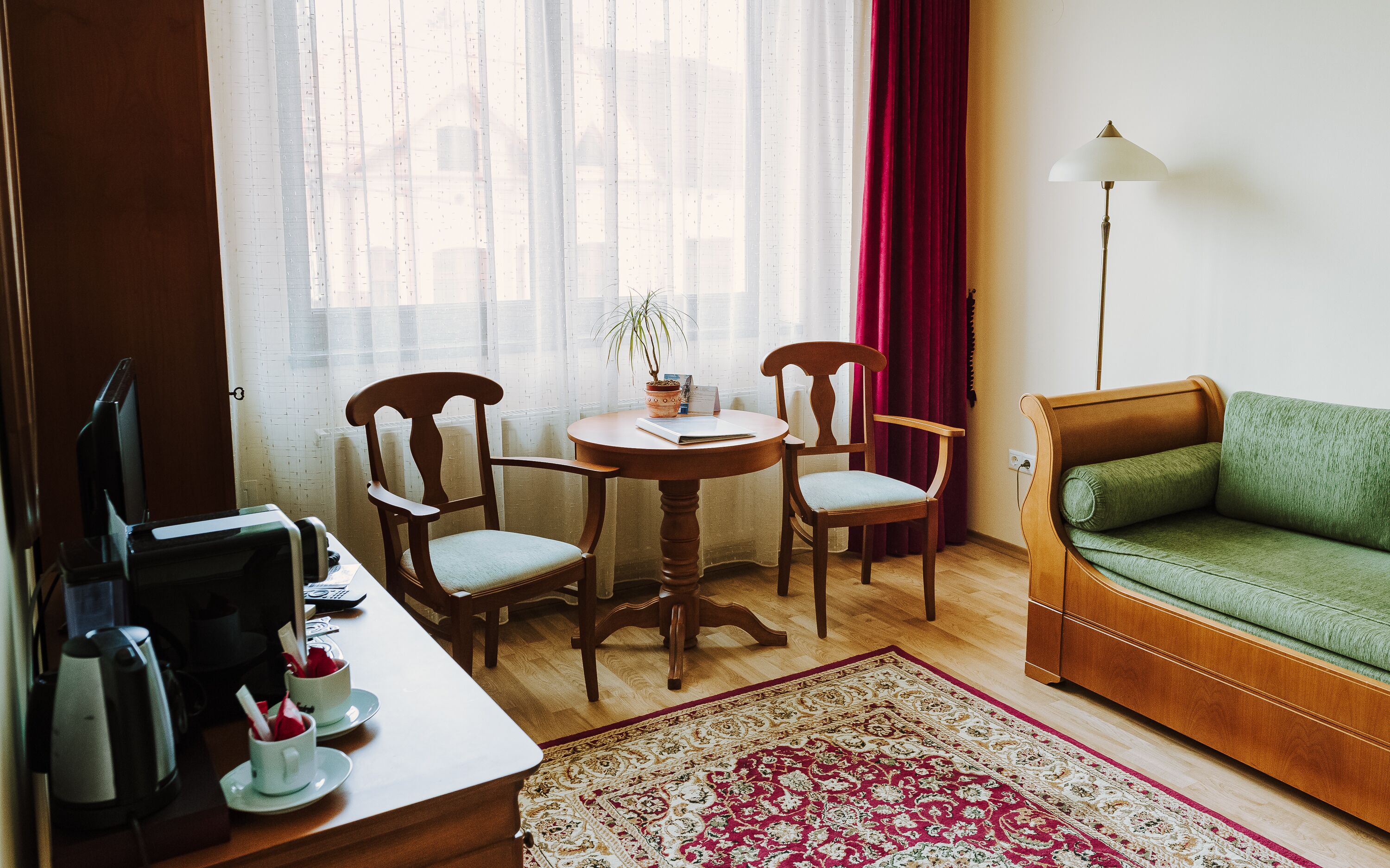 Una habitación con un escritorio de madera, dos sillas, un sofá verde, una lámpara y una ventana con cortinas translúcidas.