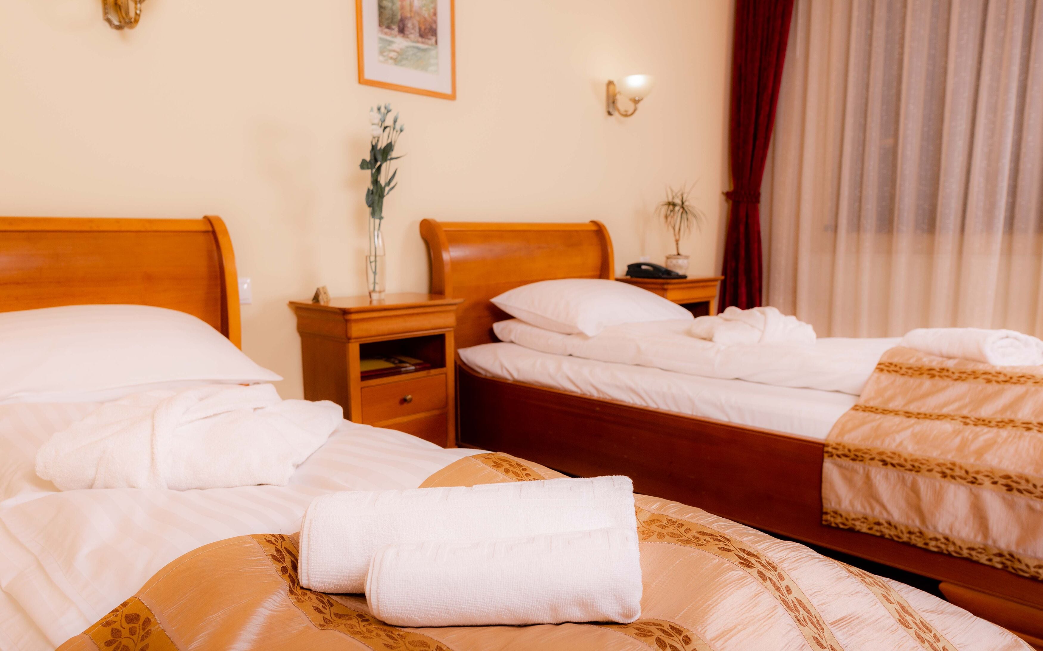 Quarto de hotel com duas camas, mesa de cabeceira, abajur, vaso com flores e janela com cortinas.