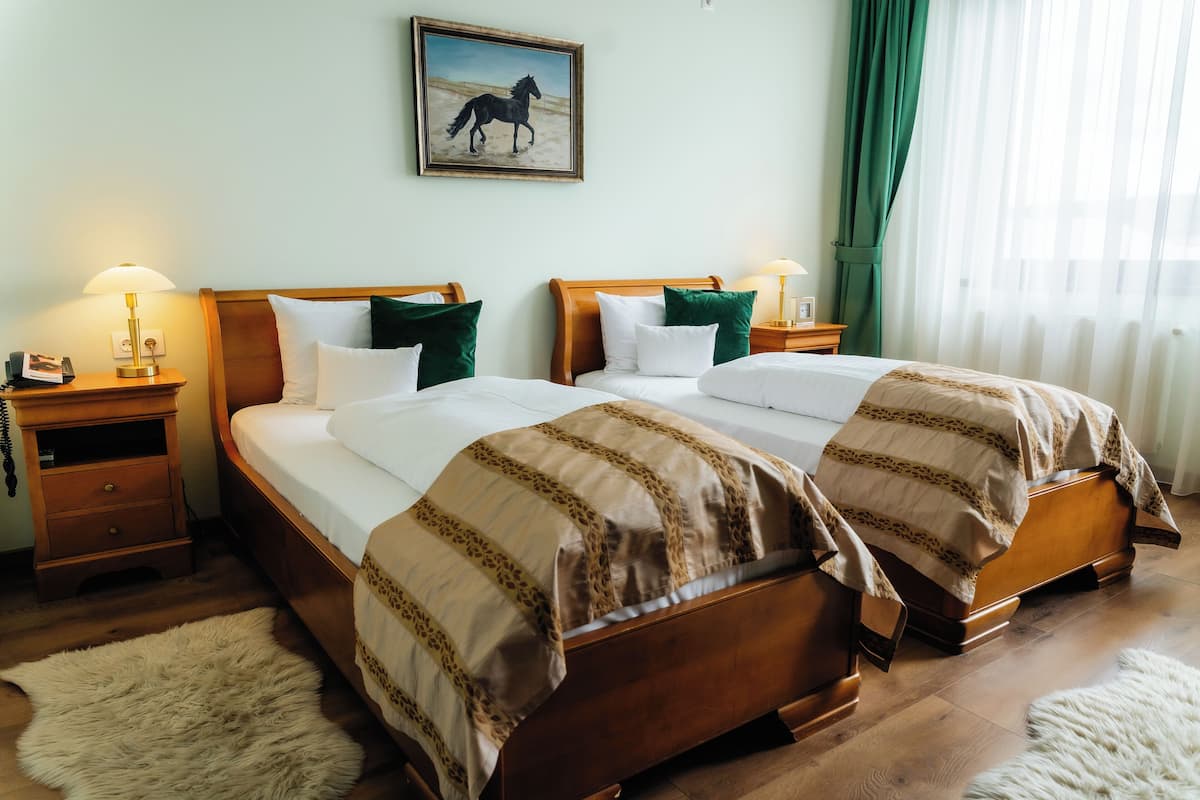 A double bed with a patterned bedspread, a nightstand with a lamp, a framed picture on the wall, and a window with curtains.