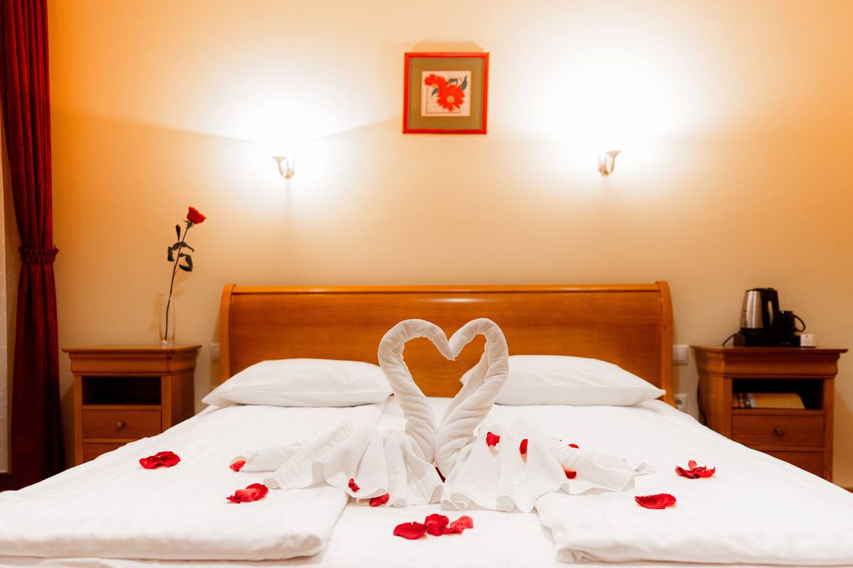 A hotel room with a neatly made bed, a heart-shaped towel arrangement, rose petals scattered on the bed, a vase with a single red rose, and a framed picture on the wall.