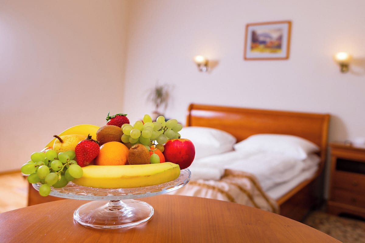 A fruit bowl on a table in a hotel room with a bed and nightstands in the background.