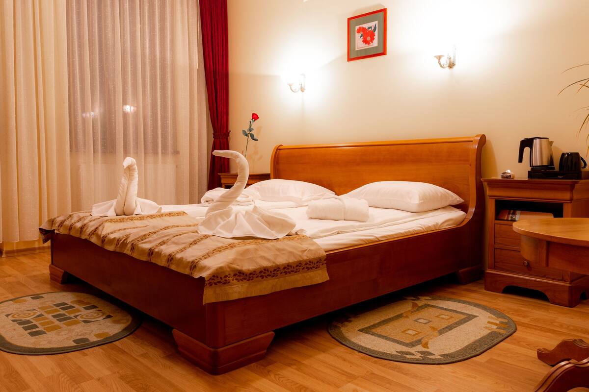 A neatly made bed with white linens and towels, a wooden headboard, a bedside table with a kettle and a book, and a red rose on the window sill.