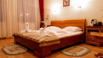 Cama bem arrumada com lençóis e toalhas brancos, cabeceira de madeira, mesa de cabeceira com uma chaleira e um livro, e uma rosa vermelha no parapeito da janela.