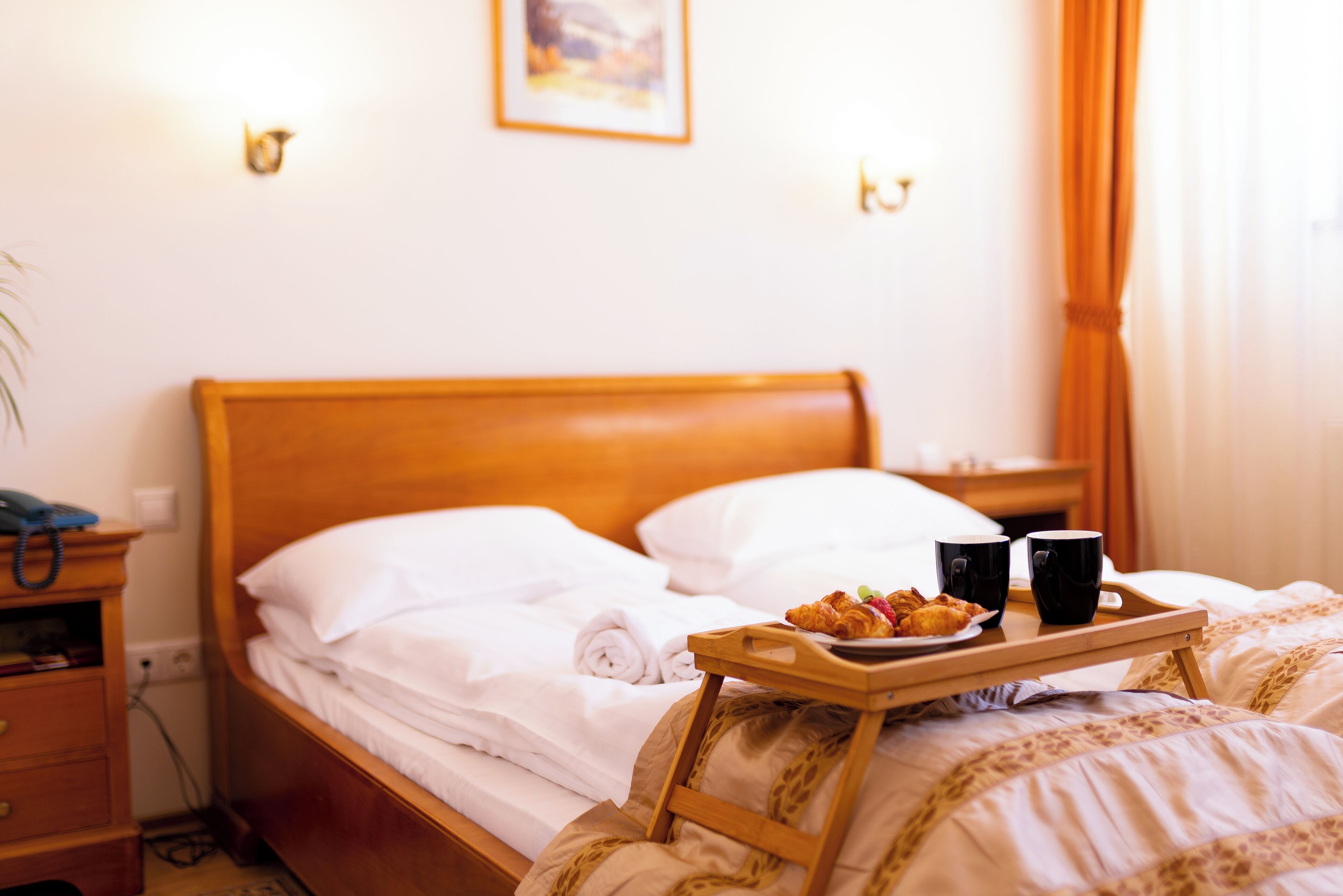 A neatly made bed with white linens, a tray with croissants and coffee cups, and a wooden nightstand with a telephone.