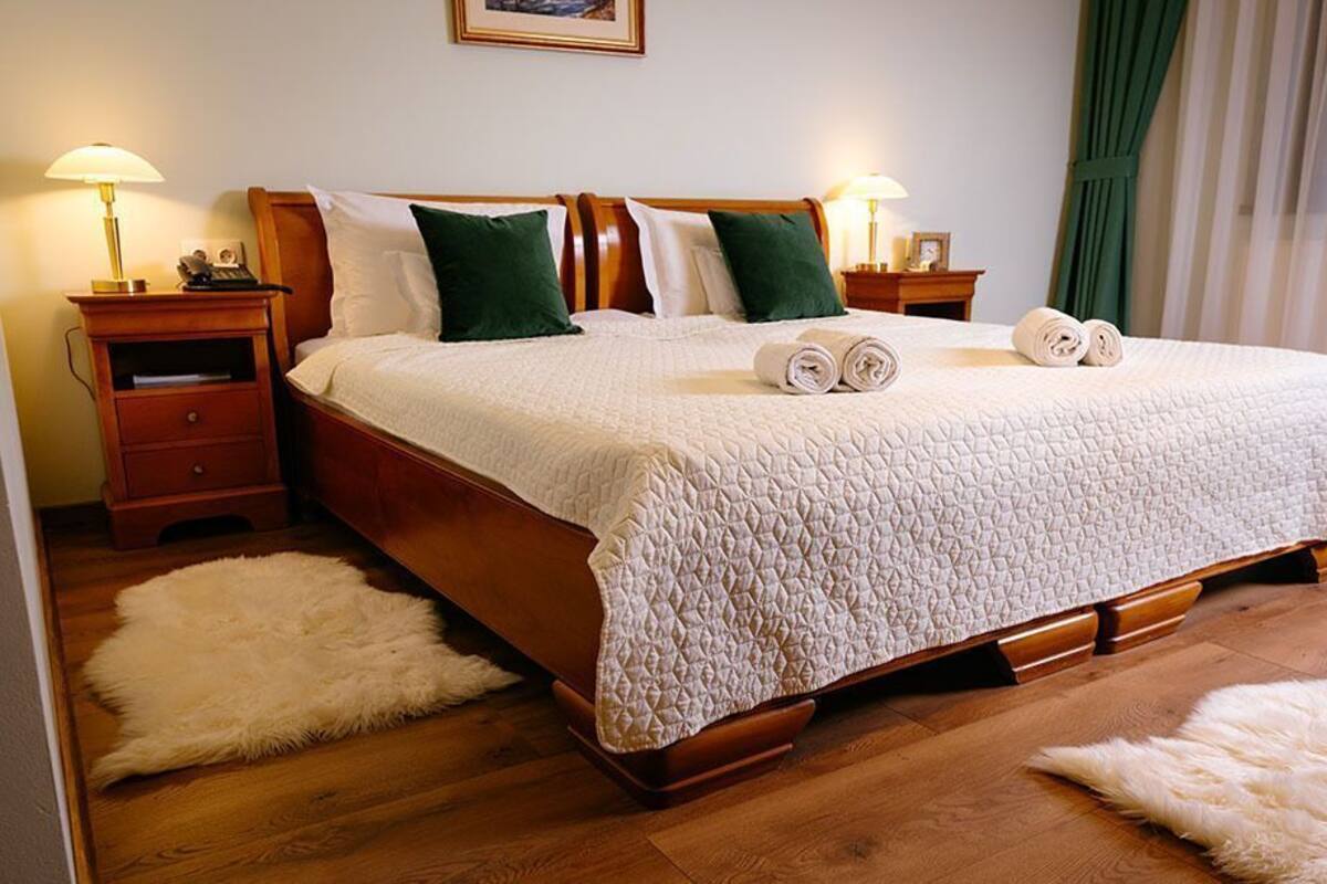 A neatly made bed with a quilted cover, two green pillows, and rolled towels on the bed. There are two bedside tables with lamps, a wooden headboard, and a fluffy rug on the floor.