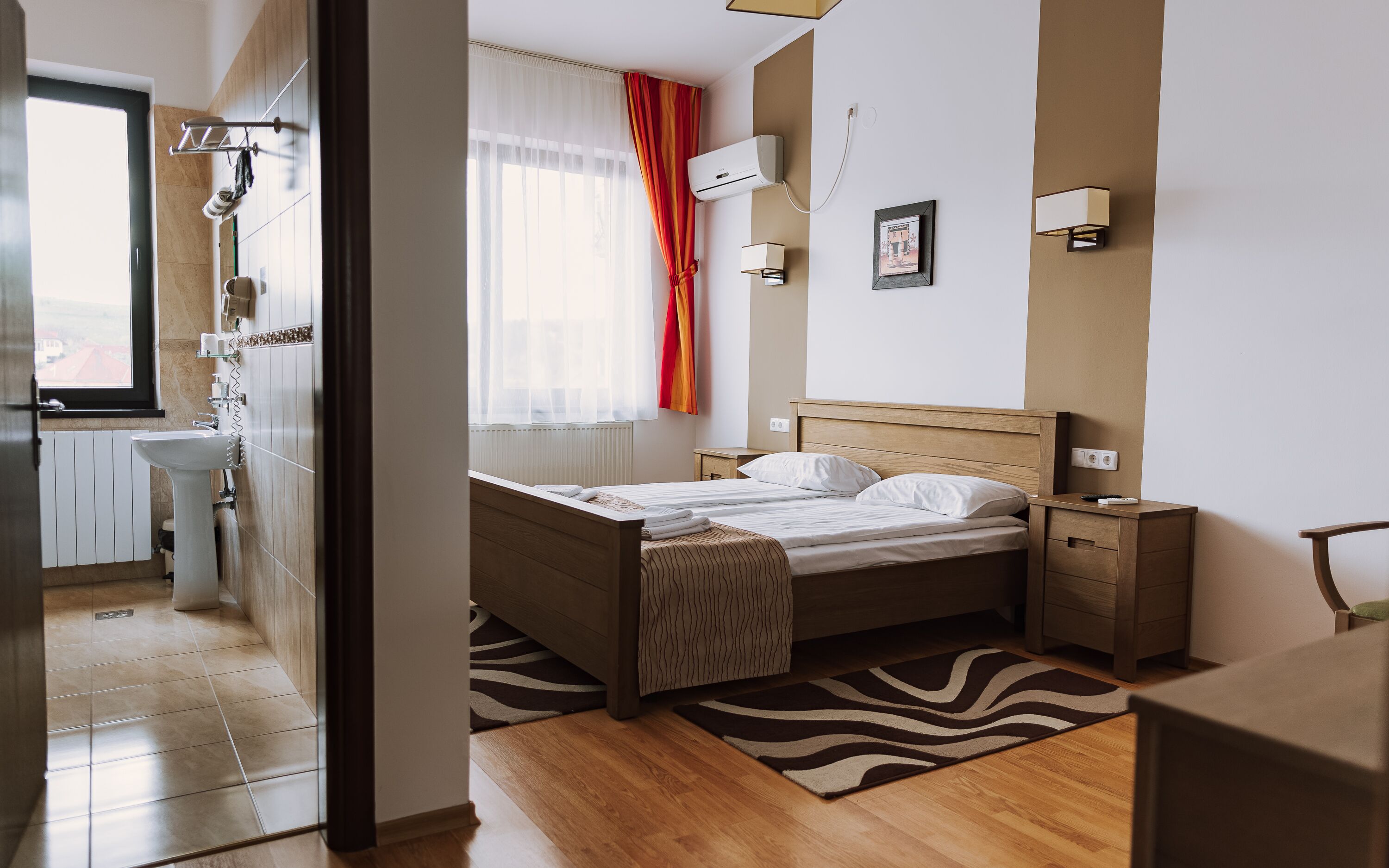 Une chambre d’hôtel moderne avec un lit en bois, une table de chevet, une commode, une fenêtre avec des rideaux, un radiateur et un climatiseur fixé au mur.