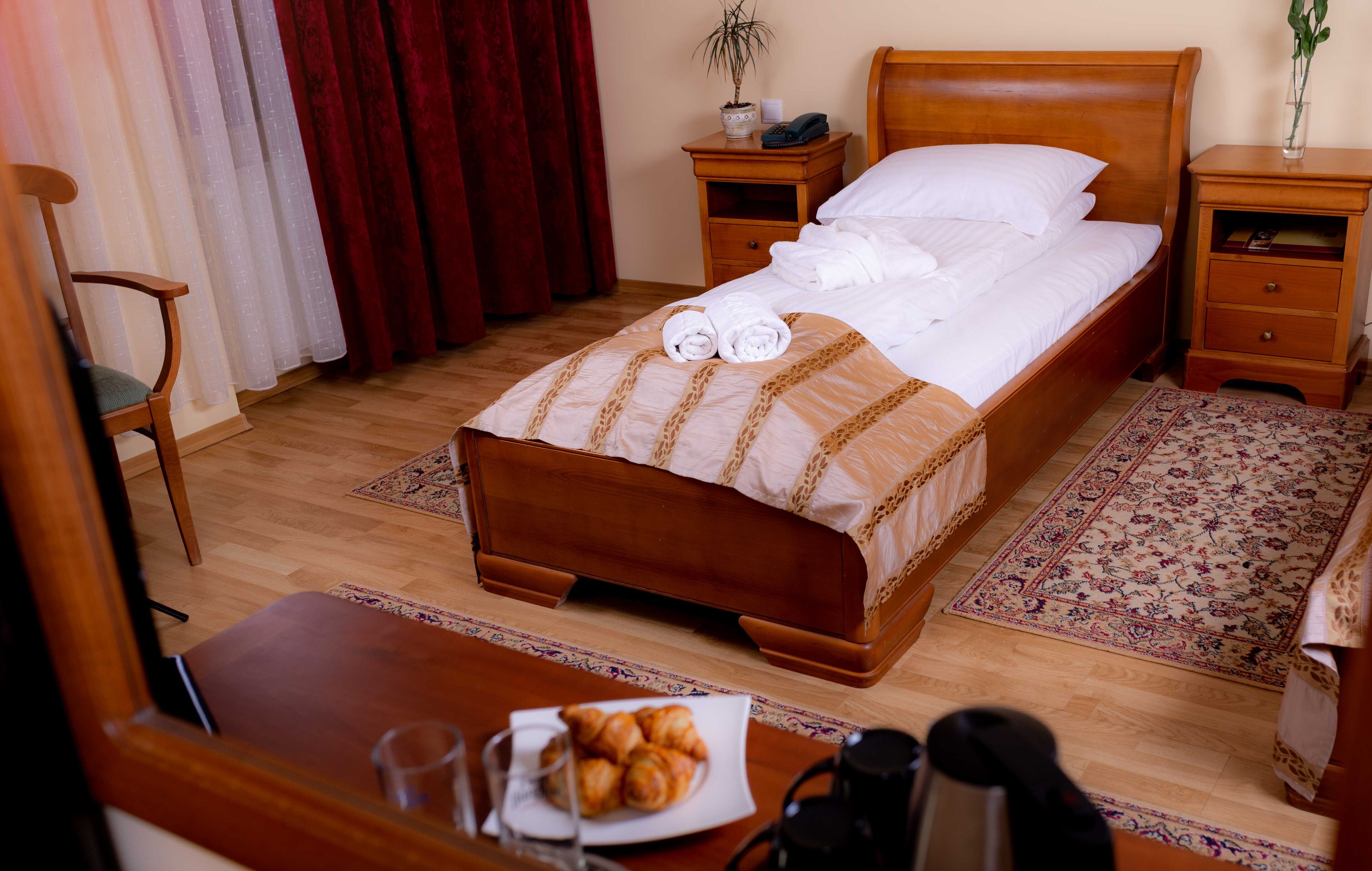 Una cama bien tendida con toallas, una mesita de noche de madera con una planta, una pequeña mesa con croissants y una cafetera, y una silla.