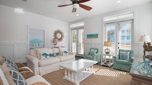 A spacious living room with a white ceiling fan, a glass coffee table, and a large window providing natural light.