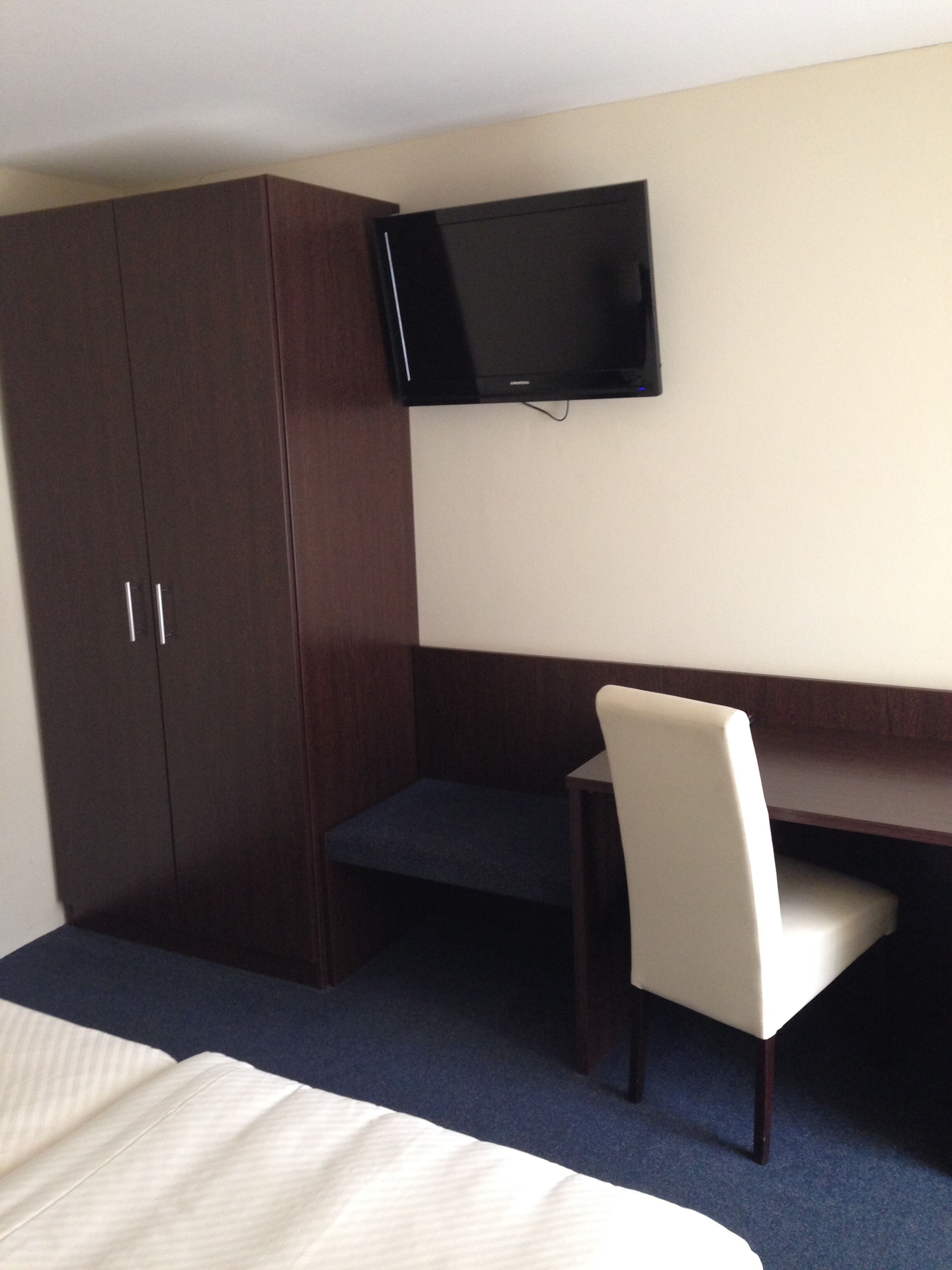 A hotel room with a bed, a desk, a chair, a television mounted on the wall, and a wardrobe.