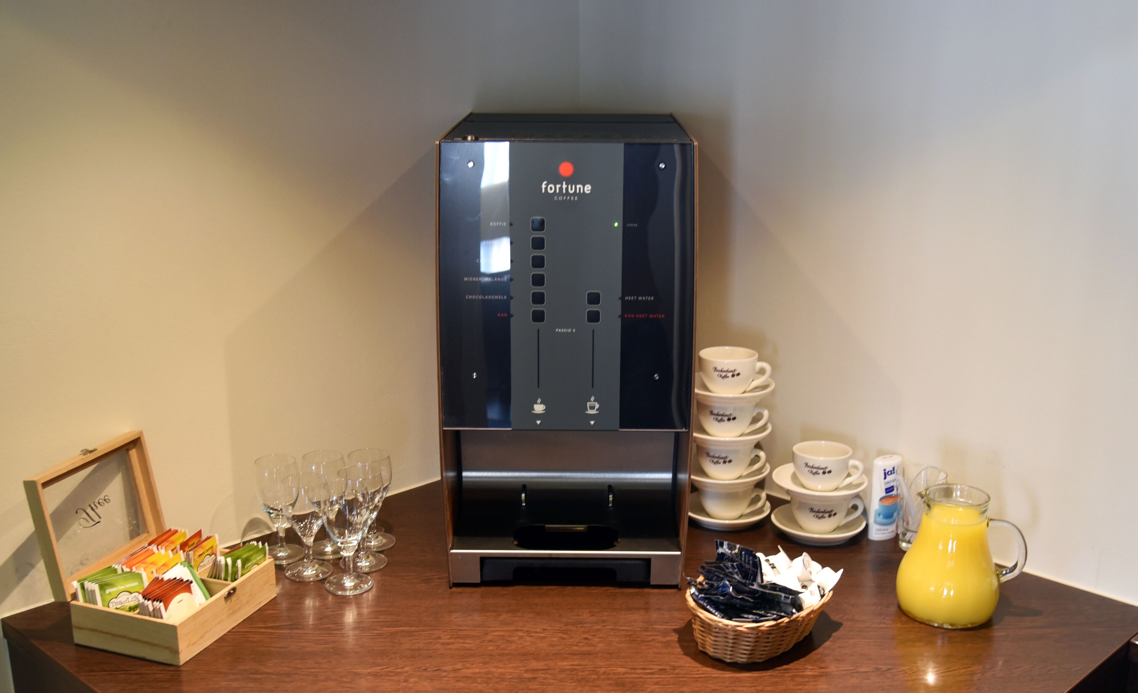 Una máquina de café con varias opciones, una pila de vasos desechables, una jarra de vidrio con jugo de naranja y una cesta de cápsulas de café.