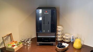 Una máquina de café con varias opciones, una pila de vasos desechables, una jarra de vidrio con jugo de naranja y una cesta de cápsulas de café.