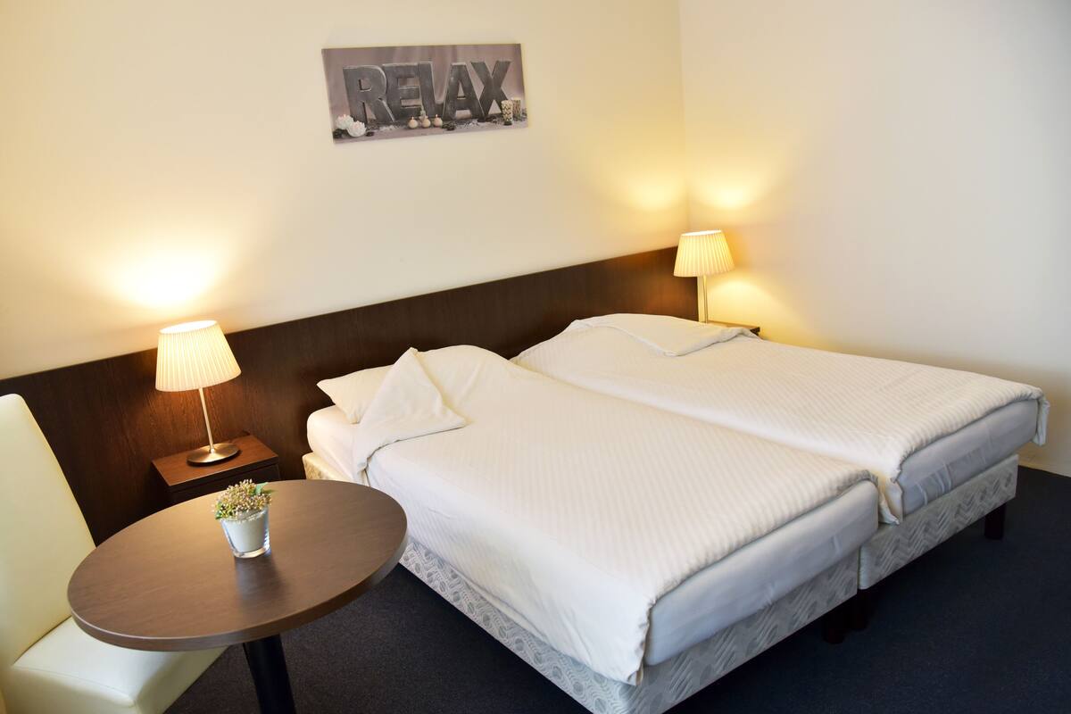 A hotel room with two beds, a small table with a lamp, a potted plant, and a 'RELAX' sign on the wall.