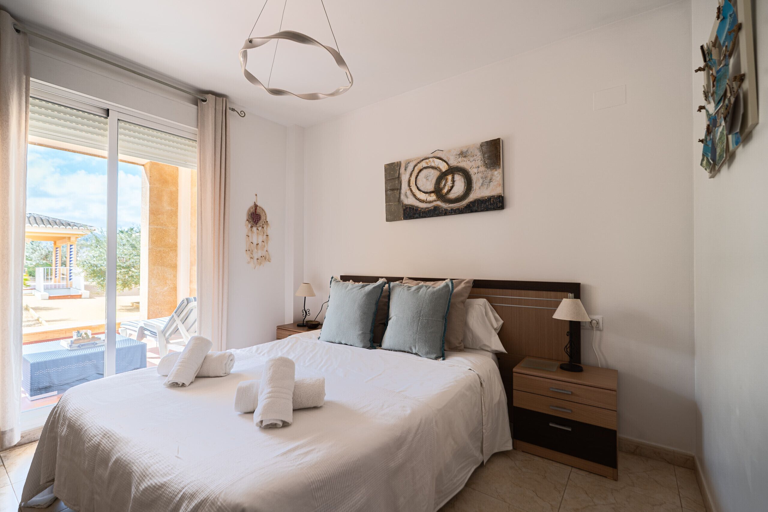 A hotel room with a bed, a nightstand, a lamp, a painting, and a view through a sliding door.